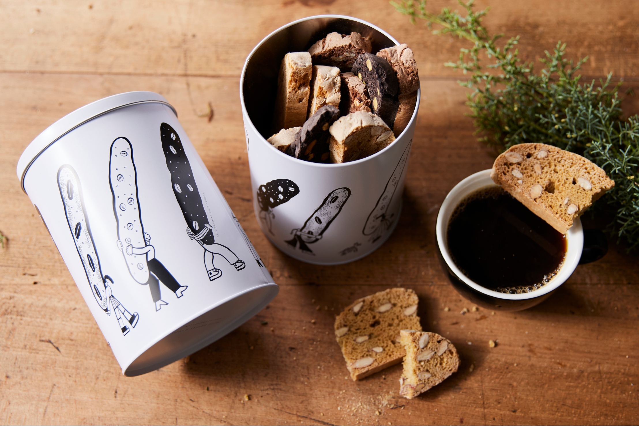 two cylindrical tins of biscotti, with images of people holding massive biscotti. A broken biscotti sits on top of a small cup of black coffee.